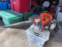 Side angled view of orange and gray Husqvarna leaf blower placed on translucent plastic container with green storage bins in background.