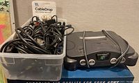 Nintendo 64 console alongside clear plastic box with tangled cables and cable clips inside.
