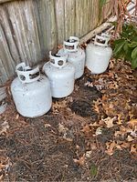 Four white propane tanks outdoors on dirt ground with dry leaves around, showing wear and dirt on tanks