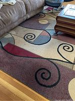 View of the rug showing broad swirls, shapes, and earth tone colors under a brown couch and near a wood floor.