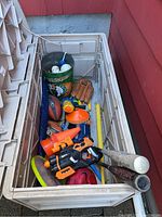 View inside plastic storage box showing sports equipment and toys including baseball bats, footballs, wiffle balls, toy guns, pylons, Boston Celtics metal bucket, kite, and soccer ball.