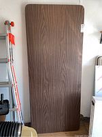 Full view of the rectangular folding table standing upright showing the brown woodgrain laminate surface.