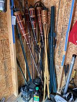 Full view of four tiki torches, green metal posts, and two solar bug zapper lights on floor in a wooden corner area.