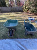 Wide view of two green plastic wheelbarrows on grass in backyard with fence and trees in the background