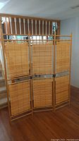 Front view of entire 3 panel wooden room divider with bamboo slat panels, standing folded upright on hardwood floor.