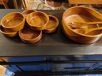 Nine-piece wooden salad bowl set with large bowl, large wooden fork and spoon, and six smaller wooden bowls arranged on a dark tabletop