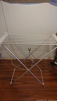 Foldable electric drying rack shown fully assembled and standing on wooden floor, with visible crossed metal legs and electrical cord.