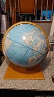 Front and side views of a large world globe showing continents including Europe, Africa, South America, and parts of Asia with blue ocean background and gold metal axis frame mounted on a wooden base.