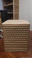 Front view of a square wicker laundry basket with a beige cushioned lid on top, showing the woven wicker details and gold tones.