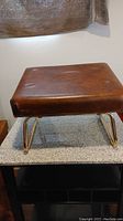Front view of brown leather cushioned foot stool with brass metal legs showing top surface and button tufting.
