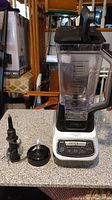 Full view of the blender with blending jar on base, two different blade assemblies beside it on counter.