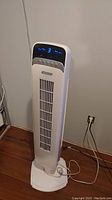 Full view of white oscillating tower fan plugged in and working showing blue digital display