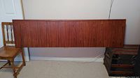 Full view of teak wood headboard, displayed against a wall, showing shape and dimensions with chair and trunk nearby for scale.