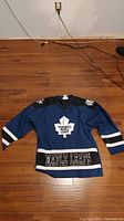 Full front view of the blue Toronto Maple Leafs youth hockey jersey on wooden floor showing graphics and color scheme.