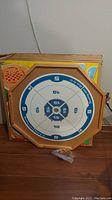 Octagonal double-sided wooden game board, Crokinole side visible with blue and white scoring zones, placed against a box.