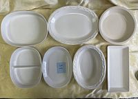 Top view of seven white ceramic serving dishes arranged on fabric background, showing variety of shapes and sizes.
