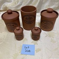 Set of six reddish-brown pottery canisters labeled sugar, salt, oregano, basil, and kitchen utensils with lids, displayed on a fabric background.