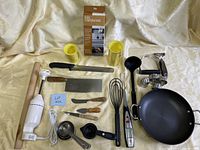 Full view of assorted kitchen tools arranged on a surface including knives, measuring cups, hand blender, ladle, whisk, peeler, thermometer, and garlic press