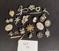 Photo of a collection of 22 assorted decorative pins and brooches arranged on a black surface with a note labeled 'Lot 39'. The pins include designs such as a faux pearl strawberry, rhinestone bow, cameo, floral motifs, rhinestone heart, enamel oval, and a rabbit figure.