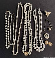Top-down view of five pearl necklaces laid out alongside assorted jewelry items. Items include Monet earrings, various brooches, pins, and a butterfly brooch.