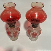 Both vintage kerosene lamps shown together, displaying red glass chimneys with scalloped tops and clear glass bases with red drop-shaped accents.