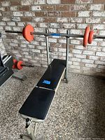 Front view of bench press with black padded cushions on beige metal frame and barbell rack with orange plates.
