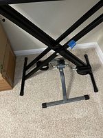 Black metal keyboard stand with X-frame shape, adjustable height mechanism, and stabilizer support at the bottom, standing on carpeted floor.