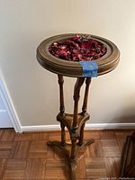 Overall view of wooden plant stand with round top filled with red dried potpourri, placed against a wall with parquet flooring.