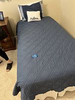 Twin bed dressed with a dark blue quilted comforter and pillows including a small 'relax' decorative pillow. Bed skirt is white. The bed is positioned next to a wooden nightstand.