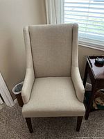 Front view of beige upholstered armchair showing high padded backrest, cushioned seat, flared arms, and dark wooden legs.