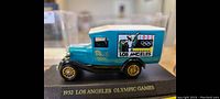 Front angle of teal diecast model car showing detailed side graphics including Olympic rings and mascot, with gold wheels on black base.