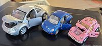 Three VW Beetle diecast model cars (silver, blue, pink) front angle showing doors and hoods open revealing interior and engine details.