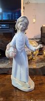 Front view of a vintage ceramic statue of a girl with a basket, glossy white finish, detailed face and dress buttons.