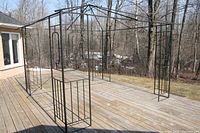 Photo showing entire metal gazebo frame standing on wooden deck outdoors, black lattice design on legs, no cover on frame.