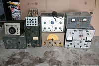 Full lot overview showing nine vintage electronics devices with various knobs, meters, and tubes, laid out on a concrete floor.