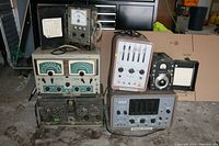 Six pieces of vintage electronic test equipment arranged on a floor, including units with analog meters, knobs, and switches. Various brand names visible.