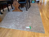 Corner view of the grey area rug placed under a wooden dining table with black chairs. Shows neutral grey plush texture and finished edges.