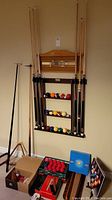 Three-shelf wooden rack mounted on wall holding cues and colored billiard balls with scoreboard above