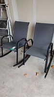 Two black metal frame patio rocking chairs with gray mesh seats and backs, shown side by side in a garage setting.
