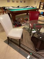 Two cream-coloured faux leather chairs and two red faux leather chairs arranged near a glass dining table and pool table in a basement setting. The broken leg is not clearly visible but noted by seller.