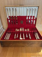 Cutlery displayed in open wooden box with red velvet and satin lining, showing knives and forks in slots and some spoons laid loose