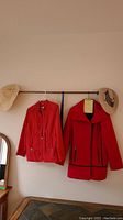 Two red ladies jackets and two sun hats hung on a wooden rod above a table, showing the full length of the jackets and positioning of hats.