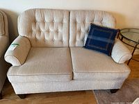 Front view of beige tufted love seat sofa with blue decorative pillow on the right side.