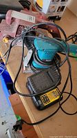 Two electric hand sanders, one Chicago Electric (reddish) and one Makita (teal), with tangled power cords and a DeWalt battery charger.