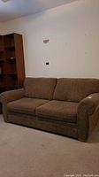 Front view of brown loveseat sofa bed with three cushions and rolled arms, placed against a wall with a bookshelf nearby.