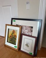 Three framed floral wall art pieces leaning against wall; one large 11x11 pink petal image, one 10x9 sunflower print, one 6x4 trillium print.