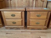 Front view of two matching solid wood bedside tables with two drawers each and brass handles, placed side by side on a wooden floor.