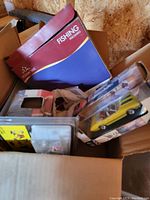 Photo of a box containing a Fishing Hollywood game, yellow die-cast car in packaging, and other assorted toys.