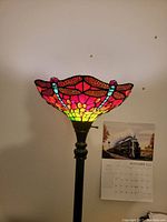 Photo showing the stained glass dragonfly lamp shade illuminated, displaying red, pink, yellow, and blue glass pieces with a scalloped edge