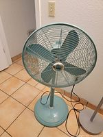 Full view of the blue vintage standing metal fan with four blades placed on a tiled floor next to a wall.